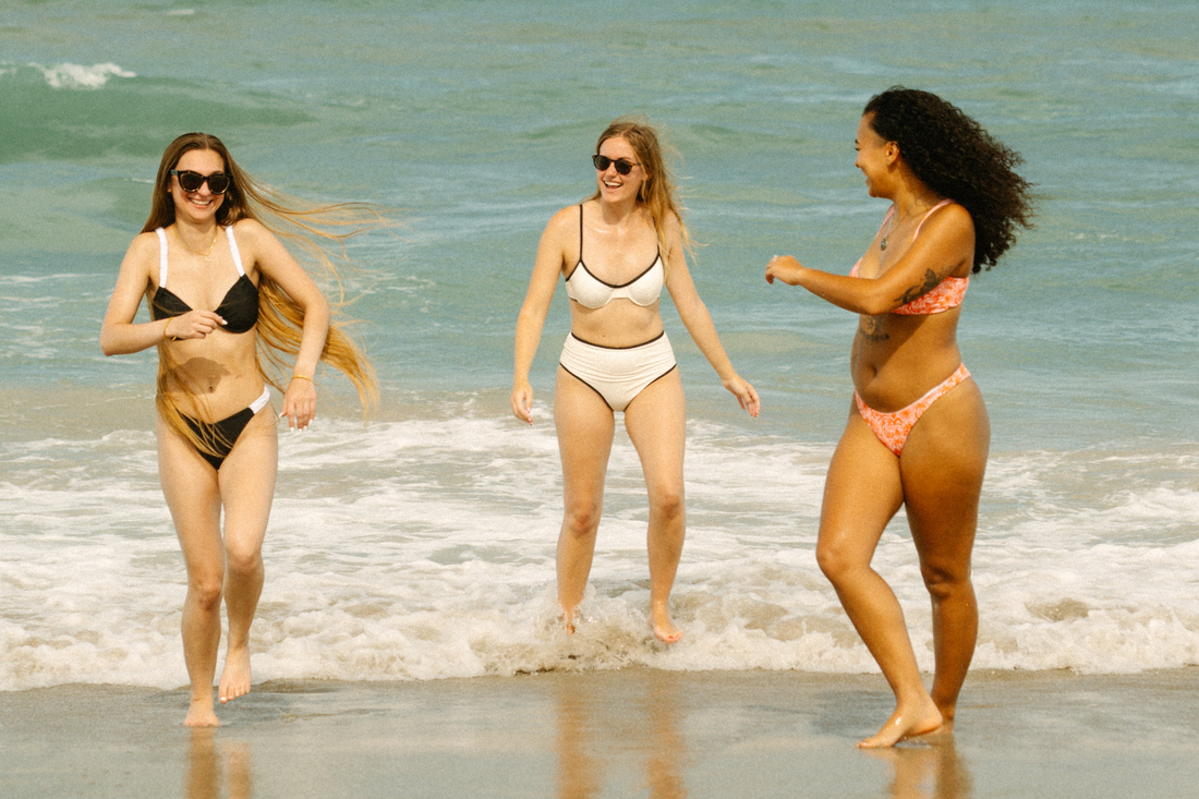 Three women in Oleka Swim bikinis run and laugh at the edge of the ocean. They are enjoying a sunny day at the beach with waves in the background