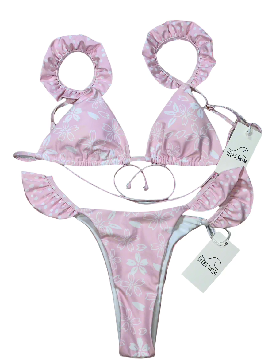 The Zuri Bikini set by Oleka Swim, displayed flat on a white background. The bikini features a light pink floral pattern with ruffled edges on the triangle top and side-tie bottom. The inner lining of the ruffles has a contrasting polka dot print. Tags labeled 'Oleka Swim' are attached to both the top and bottom.