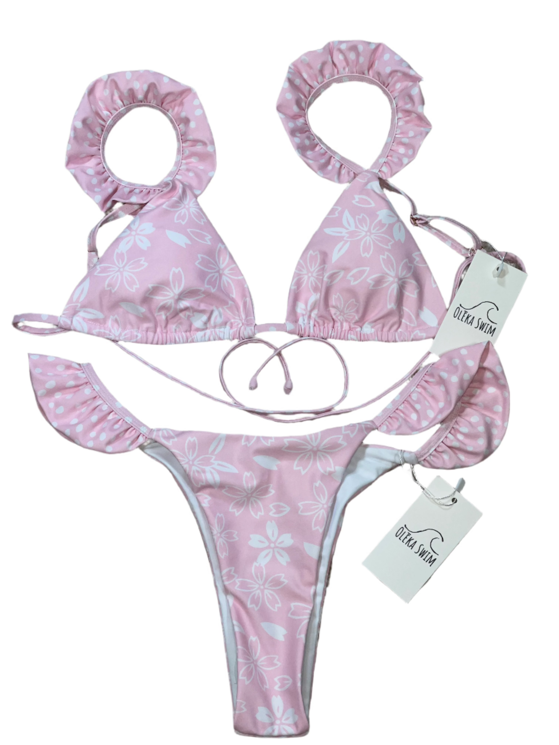 The Zuri Bikini set by Oleka Swim, displayed flat on a white background. The bikini features a light pink floral pattern with ruffled edges on the triangle top and side-tie bottom. The inner lining of the ruffles has a contrasting polka dot print. Tags labeled 'Oleka Swim' are attached to both the top and bottom.