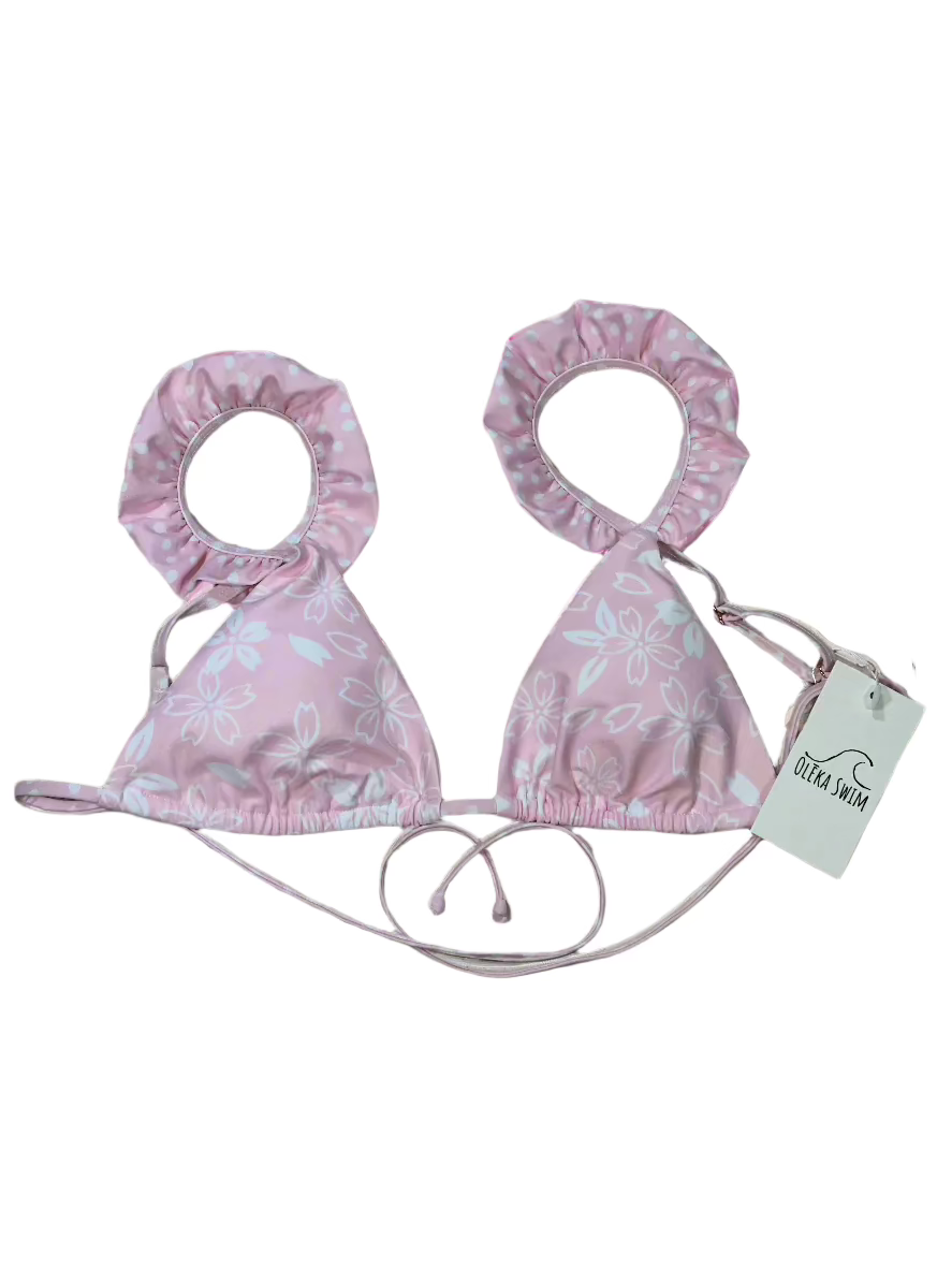 The Zuri Bikini top by Oleka Swim, displayed flat on a white background. The top is light pink with a white floral pattern, featuring ruffled edges with a contrasting polka dot print on the inner lining. A tag labeled 'Oleka Swim' is attached.