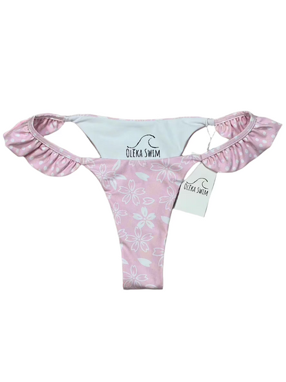 The Zuri Bikini bottom by Oleka Swim, displayed flat on a white background. The bottom is light pink with a white floral pattern, featuring ruffled sides with a contrasting polka dot print on the inner lining. A tag labeled 'Oleka Swim' is attached.