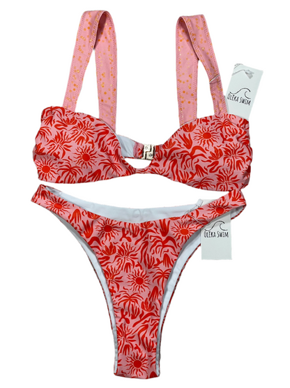 The Lola bikini set, featuring a red and pink tropical pattern with sun and palm motifs. The top has wide pink straps with a subtle dot pattern and a clasp closure at the back, while the matching bottom has a low-rise cut. The Lola bikini set is displayed flat on a white background with tags attached, labeled 'Oleka Swim.