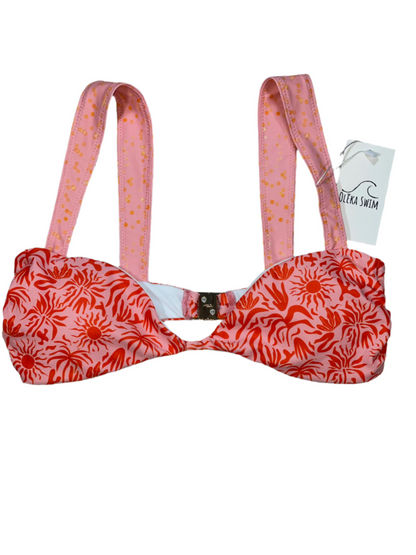 The Lola bikini top in a red and pink tropical pattern, featuring sun motifs and palm leaves. The top has wide pink straps with a subtle dot pattern and a clasp closure at the back. The bikini top is displayed flat on a white background with a tag attached, labeled 'Oleka Swim.