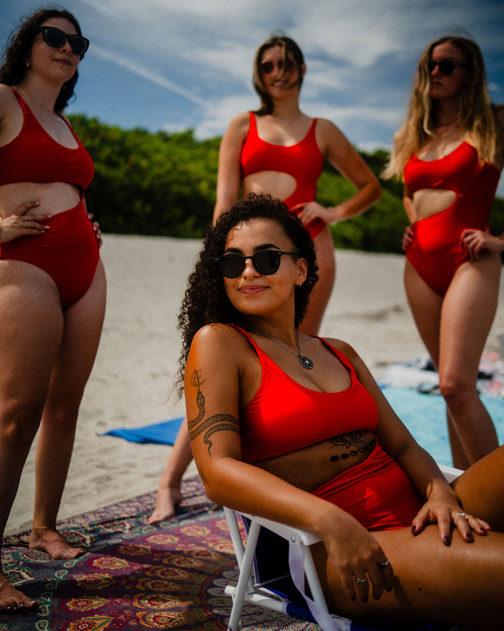 A group of four women at the beach, all wearing the Nala One Piece swimsuit from Oleka Swim in vibrant red. The women stand and sit confidently, with one seated on a beach chair in the foreground. The swimsuits feature a stylish side cutout, and the women wear sunglasses, enhancing the bold and fashionable atmosphere of the beach scene.