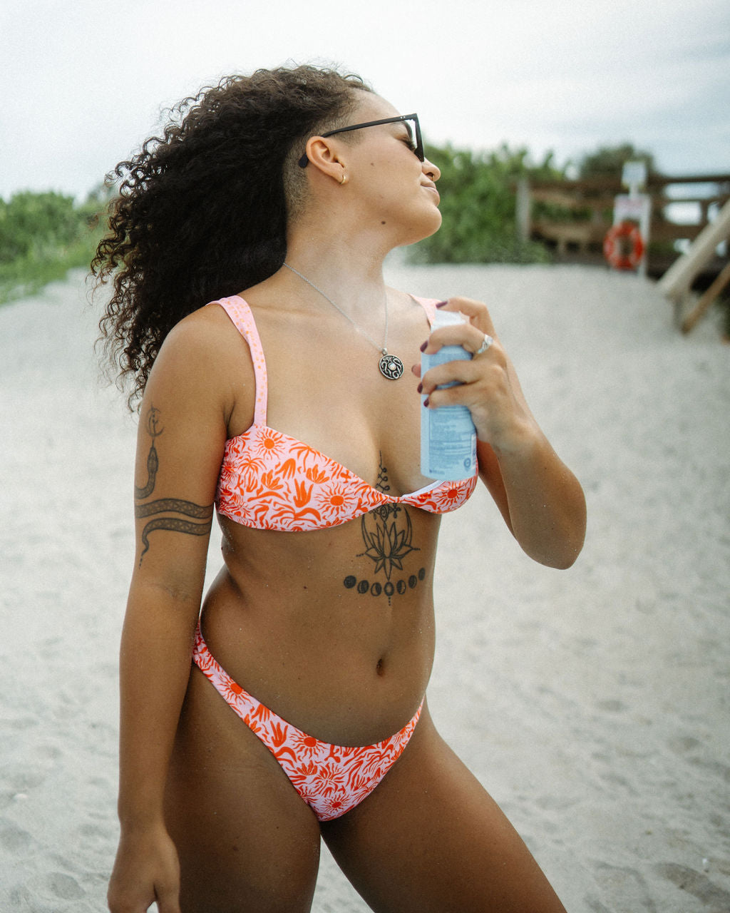 A woman wearing the Lola Bikini by Oleka Swim stands on the beach while applying sunscreen. The bikini features a vibrant red and pink tropical pattern with sun and palm motifs, with a supportive top and low-rise bottom. The woman has curly hair, wears sunglasses, and has visible tattoos on her arm and chest. She is holding a bottle of sunscreen and enjoying a day at the beach, with sand and greenery in the background.